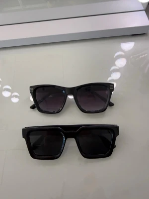 Svarta solglasögon från Ray-Ban - Klassiska svarta solglasögon från Ray-Ban med tidlös design och fyrkantig form. Bågarna är i svart plast och glasen är mörka, vilket ger en cool och stilren look. Perfekta för att lyfta vilken outfit som helst.