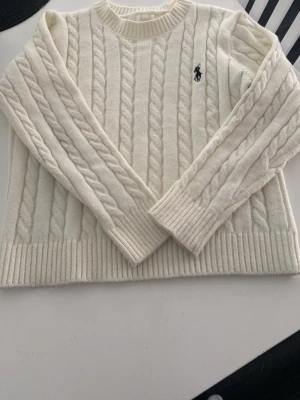 Vit kabelstickad tröja från Ralph Lauren - Kabelstickad vit tröja från Ralph Lauren med rund halsringning och ribbade muddar. Tröjan har det klassiska mörkblå logobroderiet på bröstet och är gjord i ett mjukt, tjockt material. Perfekt för kyliga dagar och enkel att styla till olika outfits. Obs! Passar inte om man är över 165cm
