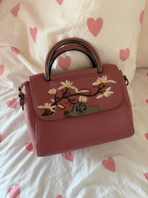 Rosa handväska med blommig brodyr - Söt handväska i rosa skinnimitation med broderade blommor i vitt, gult och brunt på locket. Väskan har ett metallhandtag i mörk silverfärg och ett vridlås framtill. Perfekt accessoar för att lyfta din outfit med en feminin touch.