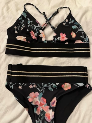 Svart blommig bikini med guldlinjer - Snygg svart bikini med blommigt mönster i rosa, vitt och grönt. Toppen har korsade axelband och bred resår med två guldiga linjer. Matchande bikiniunderdel med samma blommiga print och guldiga linjer i midjan. Perfekt för strandhäng! Har sytt in den så vet inte storlek men skulle säga att den är xs nu 