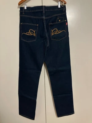 Mörkblå jeans med broderade fickor - Säljer ett par mörkblå jeans med raka ben och klassisk passform. Baksidan har ikoniska guldfärgade broderierna på bakfickorna och en liten röd etikett. Jeansen är tillverkade i kraftig denim och har fem fickor.