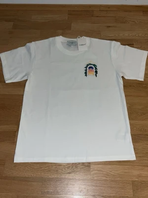 Casablanca T-shirt  - Vit t-shirt från Casablanca med färgglatt tryck av ett fönster och gröna blad på ryggen samt mindre tryck på bröstet. Klassisk passform och rund halsringning. Perfekt för dig som gillar stilrena plagg med unika detaljer.Aldrig använd!