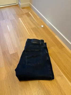 Mörkblå jeans från Nudie Jeans - Säljer ett par mörkblå jeans från Nudie Jeans med klassisk femficksmodell och vågig söm på bakfickan. Jeansen har en svart läderpatch i midjan och är tillverkade i mjuk denim. Perfekta för dig som gillar stilrena och tidlösa jeans.