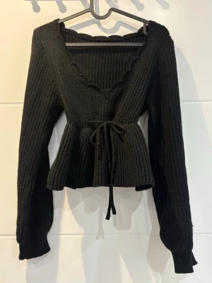 Svart stickad topp med volangkant - Säljer en svart stickad topp med djup v-ringning, volangkant och knytband i midjan. Tröjan har långa ärmar och ribbstickad struktur, samt en peplum-liknande nederdel som ger en snygg siluett. Perfekt för dig som vill ha något både gulligt och trendigt.