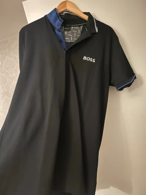 Svart pikétröja från BOSS - Svart pikétröja från BOSS med vit logga på bröstet och blå detaljer vid kragen och ärmsluten. Klassisk polokrage med knappslå och kontrastfärgade inslag. Perfekt för en stilren och avslappnad look.