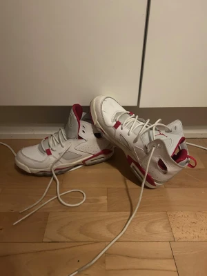 Vita Air Jordan 6 Retro sneakers - Säljer ett par vita Air Jordan 6 Retro sneakers med röda detaljer och klassisk Jumpman-logga på plösen. Skorna har högt skaft, perforerade sidor och robust sula. Materialet är en mix av läder och textil, och de har vita skosnören samt röda accenter på häl och plös.