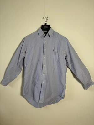 Blå skjorta från Ralph Lauren - Klassisk ljusblå skjorta från Ralph Lauren med button-down krage och broderad logga på bröstet. Skjortan har lång ärm, knappar framtill och är tillverkad i mjuk bomull. Perfekt till jeans eller chinos för en clean look.