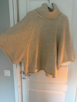 Beige stickad poncho med polokrage - Mysig beige stickad poncho med bred polokrage och vida ärmar. Perfekt att slänga på sig över en tröja för extra värme. Ponchon har en avslappnad passform och är tillverkad i ett mjukt material, oversized i modellen och är även i storlek M. Vanligtvis bär jag på storlek S men den satt även fint lite oversized