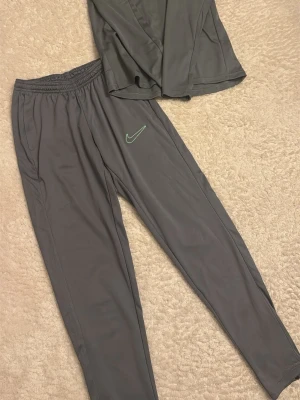 Grått träningsset från Nike - Säljer ett grått träningsset från Nike med långärmad tröja och matchande byxor. Setet har diskreta svarta detaljer och Nike-logga i ljusgrönt på både tröja och byxa. Tillverkat i ett lätt och ventilerande funktionsmaterial, perfekt för träning. Passar M oxå 