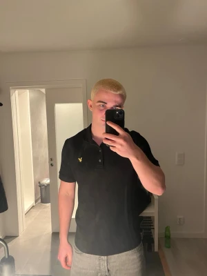Svart pikétröja från Lyle & Scott - Klassisk svart pikétröja från Lyle & Scott med korta ärmar och gul broderad logga på bröstet. Tröjan har krage och knappar framtill, perfekt för en clean och stilren look.