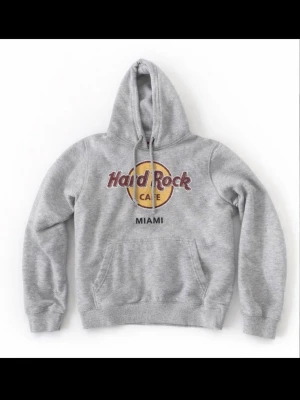 Grå Hard Rock Cafe Miami hoodie - Grå hoodie från Hard Rock Cafe med Miami-tryck framtill. Klassisk känguruficka, dragsko i huvan och ribbade muddar. Tillverkad i 80% bomull och 20% polyester för en mjuk och bekväm känsla. Perfekt för dig som gillar ikoniska loggor och streetwear-vibe.
