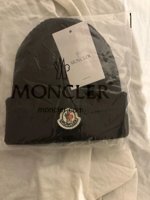 Svart ribbstickad mössa från Moncler - Snygg svart ribbstickad mössa från Moncler med uppvikt kant och klassisk Moncler-logga framtill. Perfekt accessoar för kalla dagar och ger en clean look till din outfit. Märketikett medföljer och materialet känns mjukt och värmande.