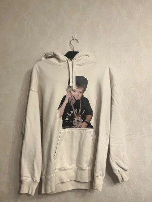 Beige hoodie med tryck från H&M - Snygg beige hoodie från H&M med stor ficka framtill och coolt tryck av en person med smycken på bröstet. Hoodien har dragsko i huvan och ribbade muddar vid ärmar och nederkant. Perfekt för dig som gillar streetwear och vill sticka ut lite extra.