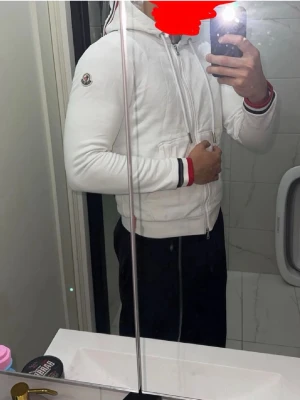 Vit Moncler hoodie med dragkedja - Säljer en fräsch vit hoodie från Moncler med dragkedja framtill och huva med snören. Tröjan har Moncler-logga på ärmen och snygga ribbade muddar med röd, vit och blå rand. Perfekt för dig som vill ha en stilren och exklusiv look. Storlek M passar S