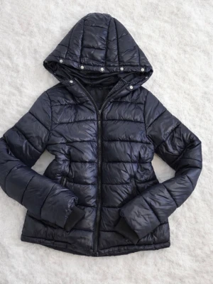 Svart pufferjacka från DeFacto - Snygg svart pufferjacka från DeFacto med huva och dragkedja framtill. Jackan har quiltad design, glansigt material och tryckknappar på huvan. Perfekt för kalla dagar och ger en modern look.