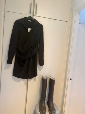 Nytt Brun skjortklänning med knyt Zara  - Stilren svart skjortklänning från Zara med lång ärm och klassisk krage. Klänningen har knappar framtill och ett snyggt knyt i midjan som ger en markerad siluett. Perfekt för dig som gillar minimalistisk och trendig stil.