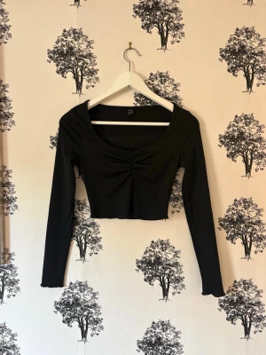 Svart croppad topp med rynk framtill - Trendig svart croppad topp med lång ärm och snygg rynkdetalj framtill. Toppen har en djup, rundad ringning och är tillverkad i ett stretchigt material som sitter tight. Perfekt för dig som gillar en stilren och enkel look.