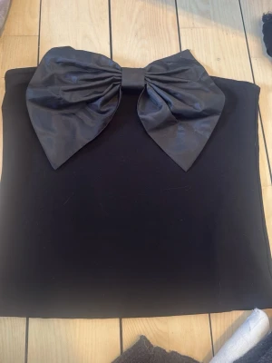 Svart axelbandslös tubtopp med rosett - Snygg svart axelbandslös tubtopp med en stor dekorativ rosett framtill. Perfekt för dig som vill sticka ut och addera en cool detalj till din outfit. Toppens material känns slätt och har en elegant finish. 700 kr ny och prislappen är kvar! Säljer för 300