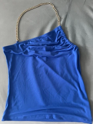 Blå  topp med guldkedja från NAKD - Snygg blå topp från NA-KD med en unik guldfärgad kedja. Toppen är ärmlös och är öppen i ryggen, perfekt för att sticka ut. Tillverkad i mjukt och stretchigt material som känns skönt mot huden.