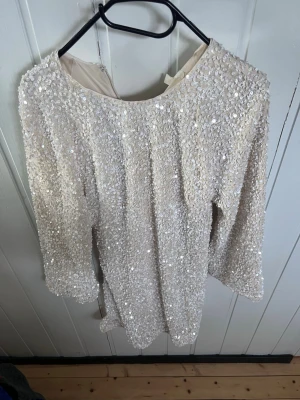 Paljettklänning i ljus champagne - Snygg kort klänning täckt av glittrande paljetter i en ljus champagnefärg. Klänningen har lång ärm och rund halsringning, perfekt för dig som vill sticka ut. Materialet känns mjukt och skimrande, och modellen är rak och enkel. (Den är sydd längst ner på höger sida, bild 5)