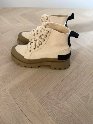 Beige chunky boots med grov sula - Säljer ett par beige boots med kraftig, grov sula i olivgrönt. Skorna har snörning framtill och svart detalj bak på hälen. Tillverkade i canvasliknande material och har en modern, chunky look som sticker ut.