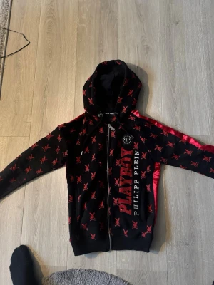Playboy x Philipp Plein hoodie s - Snygg svart hoodie från Philipp Plein i samarbete med Playboy. Hoodien har röda Playboy-kaniner över hela plagget, stor röd logga på ryggen och textdetaljer framtill. Dragkedja framtill och huva. Materialet ser ut att vara mjukt och bekvämt, perfekt för en streetwear-look.
