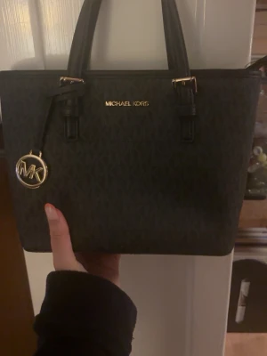 Svart handväska från Michael Kors - Snygg svart  liten handväska från Michael Kors med guldfärgade detaljer och logotyp framtill. Väskan har två handtag, strukturmönstrat konstläder och en rund MK-berlock. 