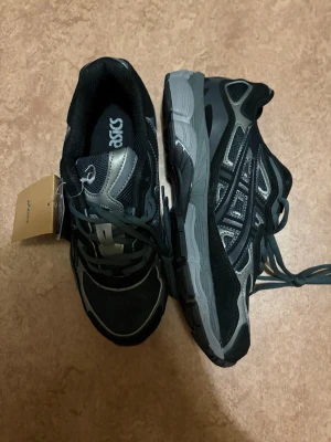 Asics gel kayano 14 skor - Snygga sneakers från Asics i svart och grått med coola detaljer och meshpaneler för bra andningsförmåga. Skorna har klassisk snörning och en robust sula som ger bra stöd. Perfekta för dig som gillar sportig stil och vill ha något bekvämt till vardags.