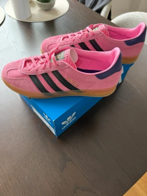 Adidas Gazelle  - Snygga Adidas Samba sneakers i rosa mocka med svarta klassiska ränder och gummisula. Skorna har lila detaljer vid hälen och guldfärgad Samba-text på sidan. Perfekta för dig som vill sticka ut med färg och stil.