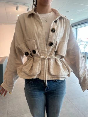 Beige overshirt med knyt och stora knappar - Säljer en beige overshirt med stora svarta knappar, två stora fickor framtill och knyt i midjan. Jackan har en avslappnad, loose passform och är tillverkad i ett mjukt bomullsliknande material. Perfekt för lager på lager och enkel att styla till jeans.