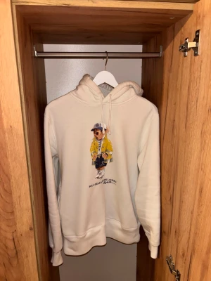 Beige hoodie Polo Ralph Lauren björn - Säljer en vit hoodie från Polo Ralph Lauren med den ikoniska björnen på bröstet, klädd i gul jacka och keps. Hoodien har dragsko i huvan och ribbade muddar vid ärmar och nederkant. Perfekt för en chill och stilren look. Tyvärr ett minimalt hål vid fickan där fram men inget märkvärdigt. 