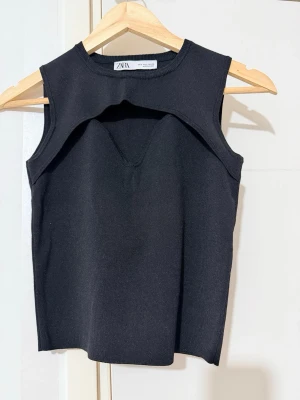 Svart ribbad ärmlös topp från Zara - Snygg svart ärmlös topp från Zara med ribbad struktur och unik detalj vid halsen. Toppen har en croppad passform och är tillverkad i ett stretchigt material som sitter bekvämt. Perfekt att styla med höga jeans eller kjol.