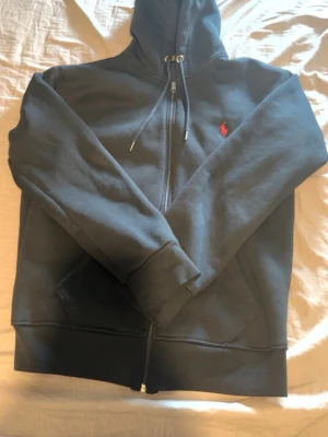 Svart hoodie från Polo Ralph Lauren - Klassisk svart hoodie med dragkedja från Polo Ralph Lauren. Har huva med dragsko, två fickor framtill och den ikoniska röda loggan broderad på bröstet. Tillverkad i mjukt bomullsmaterial som är skönt att bära. Perfekt för en avslappnad och stilren look. Priset kan diskuteras!!