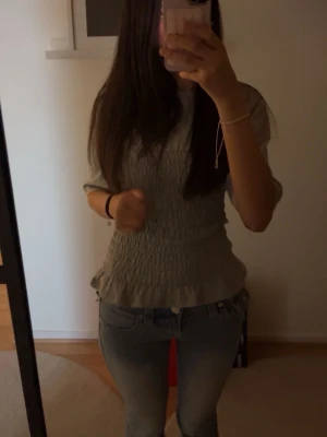Grå smockad topp med volang - Säljer en grå topp med smockad midja och volangkant nedtill. Toppen har korta ärmar och en figurnära passform som framhäver midjan. Perfekt att styla med jeans eller kjol för en trendig look.