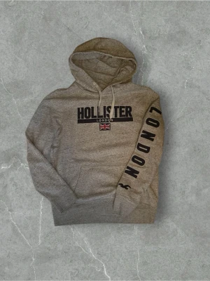 Hollister hoodie - Snygg grå hoodie från Hollister med svart text 'HOLLISTER LONDON' och brittiska flaggan på bröstet. Stor text 'LONDON' längs ena ärmen och klassisk känguruficka framtill. Mjuk bomullsblandning och justerbar huva med vita snören.