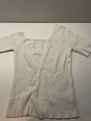 Vit ribbad topp med knappar - Trendig vit ribbad topp med asymmetrisk halsringning och dekorativa knappar framtill. Toppen har korta ärmar och är tillverkad i ett mjukt, stretchigt material som sitter snyggt på kroppen. Perfekt för dig som gillar en unik och stilren look.