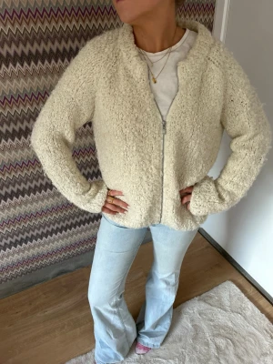 Krämvit teddykofta med dragkedja - Mysig krämvit teddykofta med dragkedja framtill och rundad hals. Jackan har en fluffig, fårskinnsinspirerad struktur och långa ärmar. Perfekt för dig som gillar en avslappnad och trendig look. Passar till jeans och sneakers för en chill stil. Passar som en M