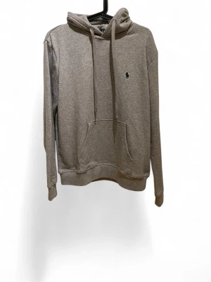 Grå hoodie från Polo Ralph Lauren S/M - Snygg grå hoodie från Polo Ralph Lauren med klassisk huva och dragsnören. Har en känguruficka framtill och den ikoniska lilla loggan broderad på bröstet. Tillverkad i mjukt bomullsmaterial som är skönt att ha på sig. Perfekt för en avslappnad och stilren look.