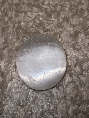 Rund selenitsten, polerad - Säljer en rund, platt och polerad selenitsten i vit genomskinlig färg. Perfekt som dekoration, för meditation eller kristallsamling. Stenen har en slät yta och är ungefär lika stor som handflatan.