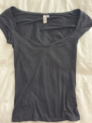 Svart topp med spetskant från Nelly - Snygg svart topp från Nelly med v-ringning och dekorativ spetskant runt halsen. Modellen är tight och har korta ärmar, perfekt att styla med jeans eller kjol. Materialet känns mjukt och följsamt.