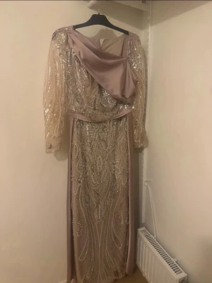 Lång glittrig champagneklänning - Elegant långklänning i champagnefärg med glittriga paljettdetaljer och transparenta långärmar. Klänningen har ett vackert mönster framtill, draperad detalj över axeln och är tillverkad i ett skimrande material som ger en lyxig känsla. Perfekt för bal eller festliga tillfällen.