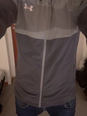 Grå vindjacka från Under Armour - Snygg tvåtonad grå vindjacka från Under Armour med hel dragkedja och huva. Jackan har en vit logga på bröstet, praktiska sidofickor med dragkedja och är tillverkad i ett lätt, vindtätt material. Perfekt för blåsiga dagar och sportiga outfits.
