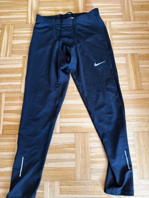 Svarta Nike Dri-FIT träningstights - Säljer ett par svarta Nike Dri-FIT träningstights med reflekterande detaljer vid benen och klassisk Nike-logga på låret. Tillverkade i ett stretchigt och snabbtorkande material som passar perfekt för löpning eller gym. Dragsko i midjan för bästa passform.
