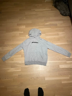 Grå hoodie från MM Sports - Snygg grå hoodie från MM Sports med svart logga på bröstet. Klassisk modell med huva och stor magficka. 