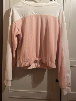 Rosa och vit sweatshirt med dragsko - Mjuk sweatshirt i ljusrosa och vitt med långa ärmar och rund hals. Tröjan har en snygg dragsko i midjan för justerbar passform och breda muddar vid ärmsluten. Perfekt för en avslappnad och trendig look.