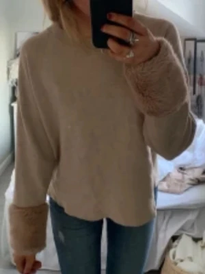 Beige stickad tröja med fuskpälsmuddar - Säljer en beige stickad tröja med breda, fluffiga fuskpälsmuddar på ärmarna. Storlek M från Holly & Whyte.