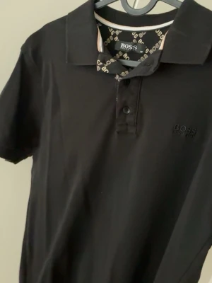 Svart polotopp från BOSS - Klassisk svart polotopp från BOSS med korta ärmar och diskret logga broderad på bröstet. Tidlös design med krage och knappar framtill. Tillverkad i mjuk bomull för en bekväm känsla. Perfekt till jeans eller chinos. Hör av dig om du har några frågor.