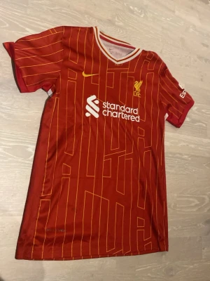 Liverpool matchtröja Nike röd - Säljer en röd Liverpool matchtröja från Nike med gula detaljer och tunna gula linjer. Tröjan har klubbmärke och sponsortryck på bröstet samt vit och gulrandig V-ringad krage. Perfekt för dig som älskar fotboll och vill stötta ditt lag.
