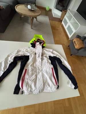 Vit vindjacka Five Seasons med huva - Snygg vit vindjacka från Five Seasons med mörkblå detaljer längs ärmarna, röda dragkedjor och en huva i neon-gult. Jackan har meshfoder och fleece vid kragen för extra komfort. Perfekt för blåsiga dagar och outdoor-aktiviteter.