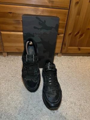 Valentino rockrunners  - Säljer ett par svarta sneakers från Valentino med mörkgrönt camomönster. Skorna har låga skaft, klassisk snörning och är tillverkade i en mix av mocka och textil. Men tyvärr lite hål i hälen efter andvändning. Snygg och stilren design med diskret logga på plösen. Perfekta för dig som gillar exklusiva skor. Pris går att diskutera.
